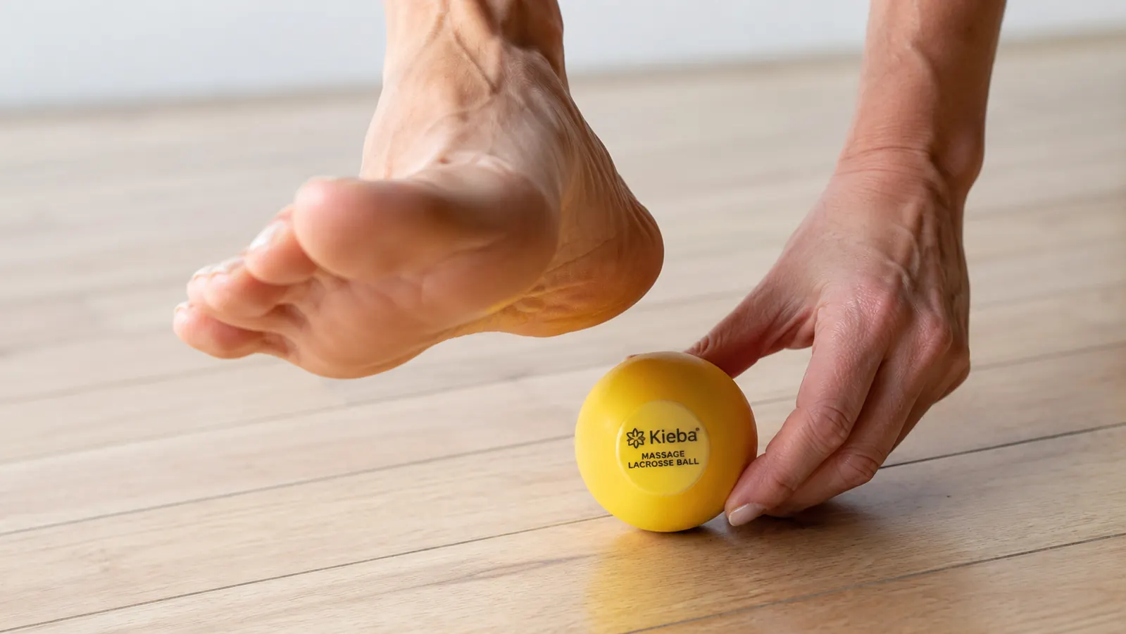 Close-up of a hand placing a Kieba lacrosse ball on a hardwood floor next to a bare foot