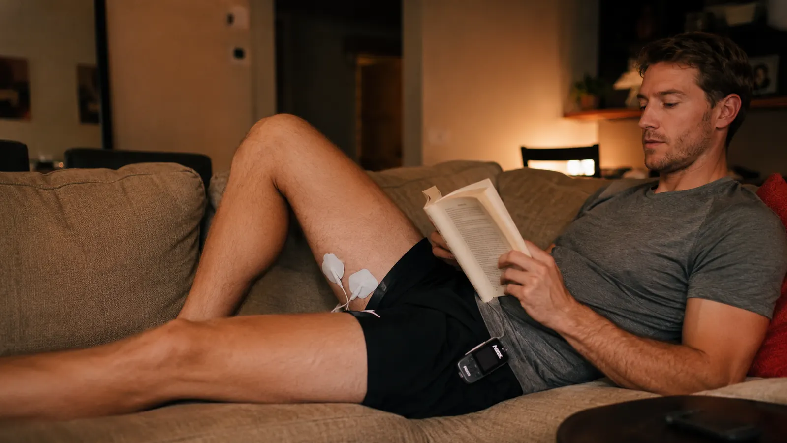 Relaxed athlete lying on a couch using a TENS unit on sore quads after a leg workout