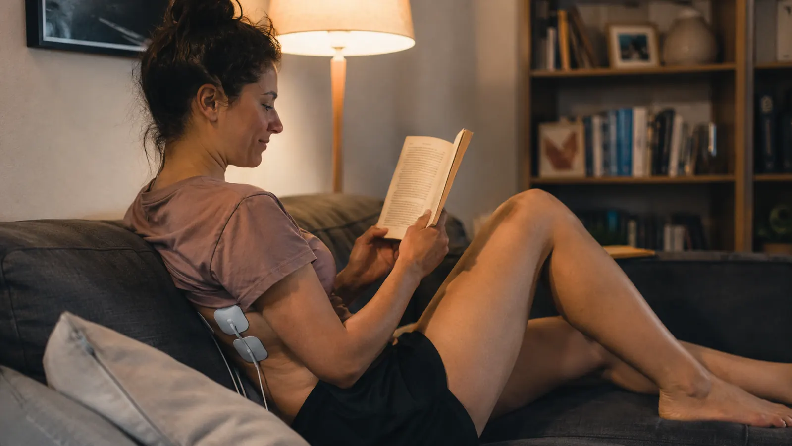 Relaxed woman sitting on a couch with TENS unit pads on her lower back, reading a book, looking comfortable